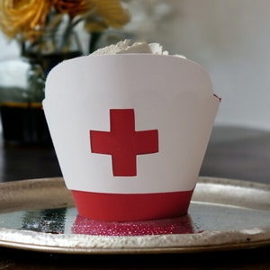 May include: A white and red cupcake wrapper shaped like a nurse's cap, with a red cross on the front. The wrapper sits on a silver tray, filled with white substance. A vase of yellow flowers is in the background.
