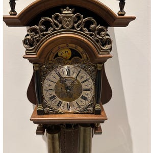Op de afbeelding: Een vintage houten wandklok met een gedetailleerde messing wijzerplaat, Romeinse cijfers en decoratieve gravures. De klok heeft twee messing gewichten die eronder hangen en sierlijke details aan de boven- en zijkanten. Een klassiek stukje woondecoratie.