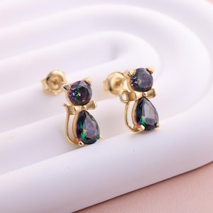 Puede incluir: Un par de pendientes de oro en forma de gato con detalles de piedras preciosas iridiscentes. Los pendientes son pequeños y delicados, perfectos para el uso diario.