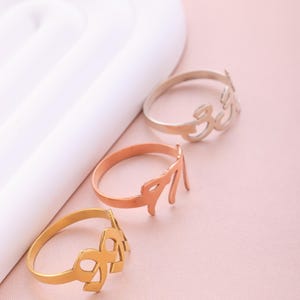 Puede incluir: Tres anillos de oro con diferentes nombres escritos en escritura &aacute;rabe. Los anillos est&aacute;n sobre una superficie rosa.