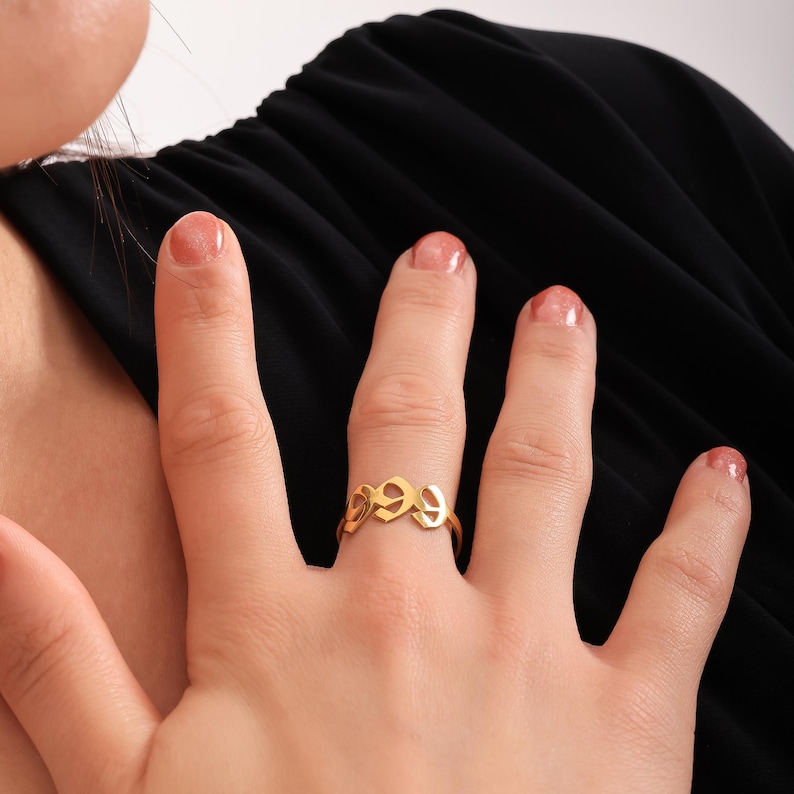 Puede incluir: Un anillo de oro con un dise&ntilde;o &uacute;nico que presenta letras entrelazadas. El anillo se lleva en el dedo de una persona con piel clara.