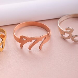 Puede incluir: Un anillo de oro rosa con un nombre escrito en cursiva. El anillo est&aacute; sobre una superficie rosa.