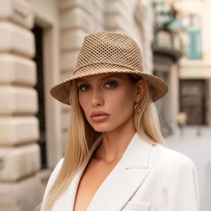 May include: A woman wearing a white blazer and a light brown woven fedora hat. The hat has a wide brim and a mesh design.