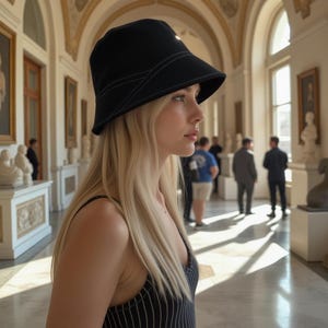 May include: A woman with long blonde hair wearing a black bucket hat with a white stitch detail and a black and white striped tank top.