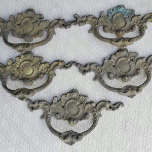 May include: Four ornate, antique-style drawer pulls with a decorative, scrolled design. The pulls are made of metal and have a tarnished, aged finish.