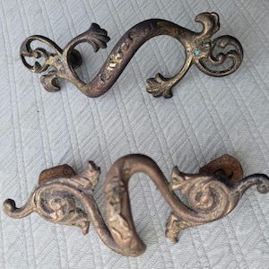 Könnte beinhalten: Zwei verzierte Metall-Schubladengriffe mit einem dekorativen, wirbelnden Design. Die Griffe sind aus dunklem, gealtertem Metall mit einer leicht angelaufenen Oberfläche.