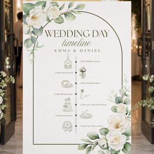 May include: A wedding day timeline sign featuring white roses and green eucalyptus leaves. The text reads Wedding Day timeline, Emma & Daniel, and lists events from 1:00pm to 11:30pm with icons for a church, drinks, breakfast, microphone, cake, food, and car.
