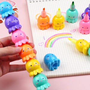 Puede incluir: Un conjunto de rotuladores de colores en forma de pulpo. Los rotuladores están conectados entre sí en una cadena, con cada pulpo de un color diferente: morado, rosa, naranja, amarillo, azul claro y azul oscuro. Los rotuladores están sobre una superficie blanca con un arcoíris dibujado en ella.