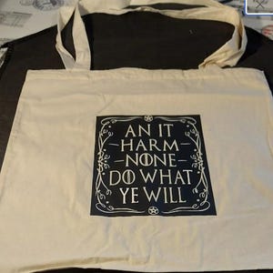 May include: A white canvas tote bag with black handles. The bag has a black square design with white text that reads "An it harm none do what ye will".