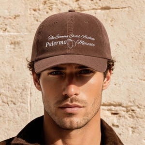 May include: A brown corduroy baseball cap featuring white embroidered text that reads The Evening Social Archive Palermo Mercato with a small line-art graphic of two lemons positioned between the words Palermo and Mercato on the front panel.