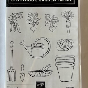 May include: A clear plastic case containing a cling stamp set titled STORYBOOK GARDEN PATCH. The set features black line art illustrations of garden items including peas, a beet, strawberries, a carrot, a watering can, a flower, a basket, and pots.