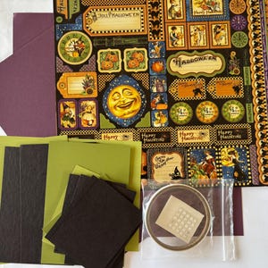 May include: A Halloween-themed craft kit featuring a large sheet of decorative paper with vintage-style illustrations of moons, pumpkins, and text, accompanied by dark purple envelopes, solid green and black cardstock, and a roll of adhesive dots.