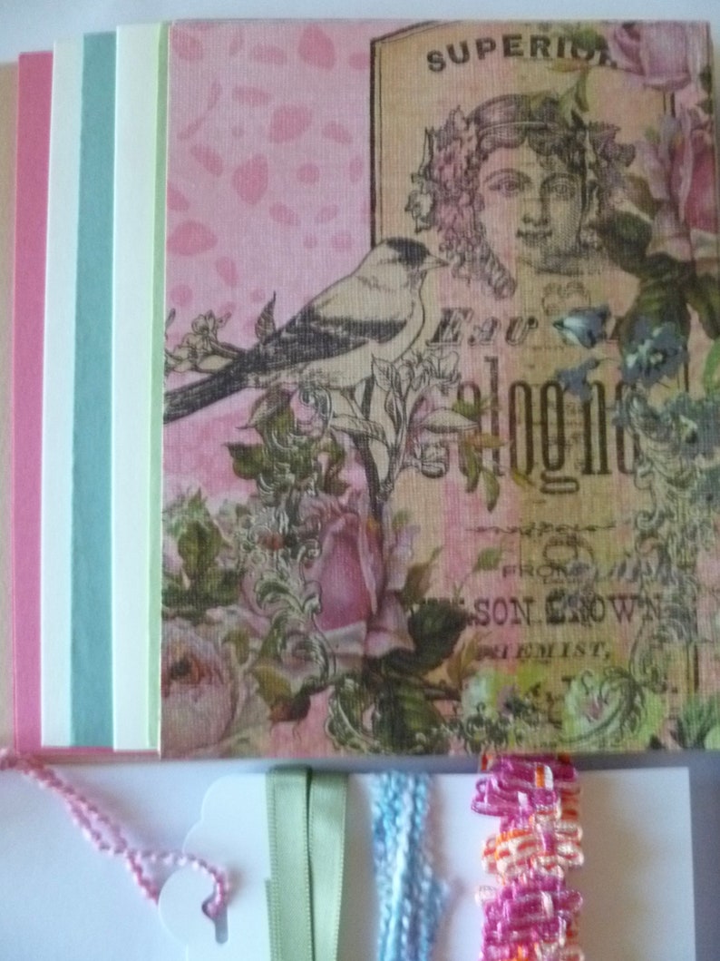 May include: A pink and white patterned paper with a bird, flowers, and the text "Superior" and "E.W. Blogg" on it. There are also several strips of paper in different colors, including pink, blue, and green, and ribbons in green, blue, and pink.
