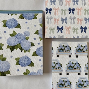 May include: Three rectangular greeting cards with distinct patterns. One features blue hydrangea clusters with green leaves, another displays a repeating grid of small bows in blue, pink, and green, and the third shows blue pickup trucks carrying hydrangeas.