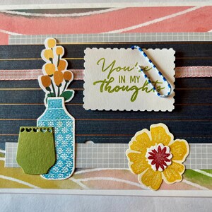 May include: A handmade greeting card with a floral theme. It features a blue vase with flowers, a yellow flower, and a paper clip. The card includes the text "You're in my thoughts."