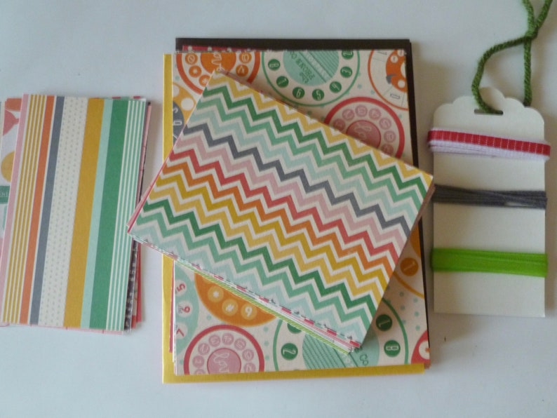 May include: A collection of patterned paper goods, including notebooks and cards. The notebooks feature a colorful chevron pattern, while the cards display a retro telephone dial design. A tag with colorful threads and ribbons is also included.