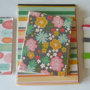 May include: A collection of stationery items, including notebooks and a tag. The notebooks feature various patterns, including floral, speech bubbles with text, and striped designs. The tag has colorful ribbons and green twine.