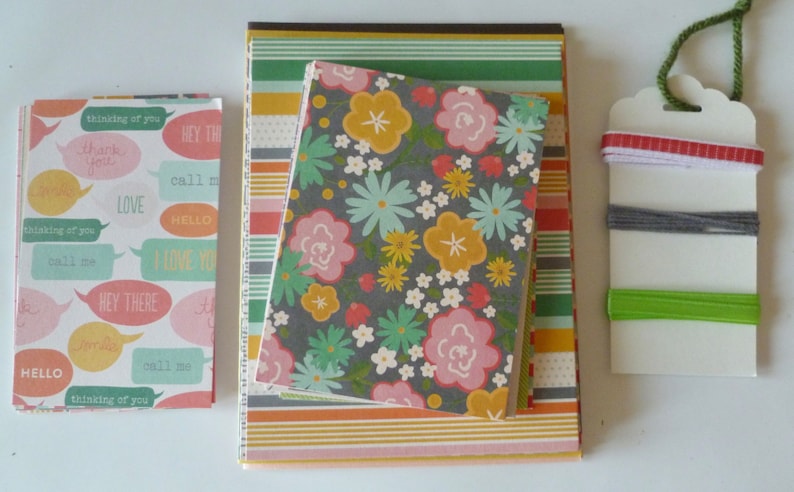 May include: A collection of stationery items, including notebooks and a tag. The notebooks feature various patterns, including floral, speech bubbles with text, and striped designs. The tag has colorful ribbons and green twine.