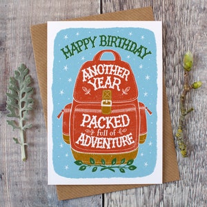 May include: A birthday card with a blue background and a red backpack with the text "Happy Birthday Another Year Packed Full of Adventure".