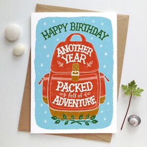 May include: A birthday card with a blue background and white stars. The card features a red and brown backpack with the text "Happy Birthday Another Year Packed full of Adventure".