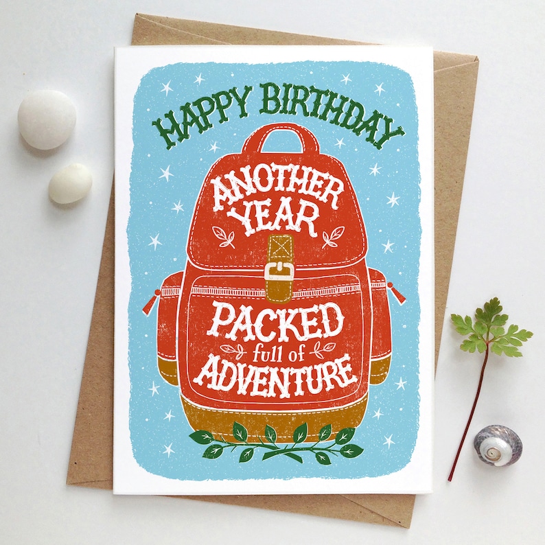 May include: A birthday card with a blue background and white stars. The card features a red and brown backpack with the text "Happy Birthday Another Year Packed full of Adventure".