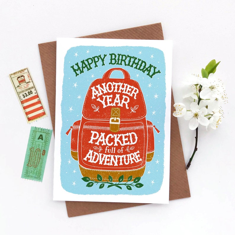May include: A blue and white birthday card with a red backpack illustration. The text on the card reads "Happy Birthday Another Year Packed Full of Adventure".
