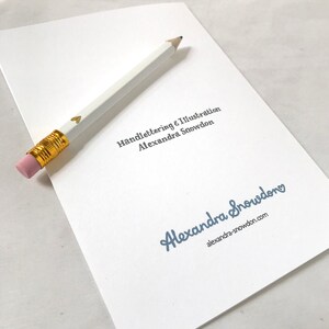 May include: A white card with the text "Handlettering & Illustration Alexandra Snowden" in gold lettering. Below the text is a blue signature that reads "Alexandra Snowden" with a small heart next to it. The text "alexandra-snowdon.com" is printed below the signature.
