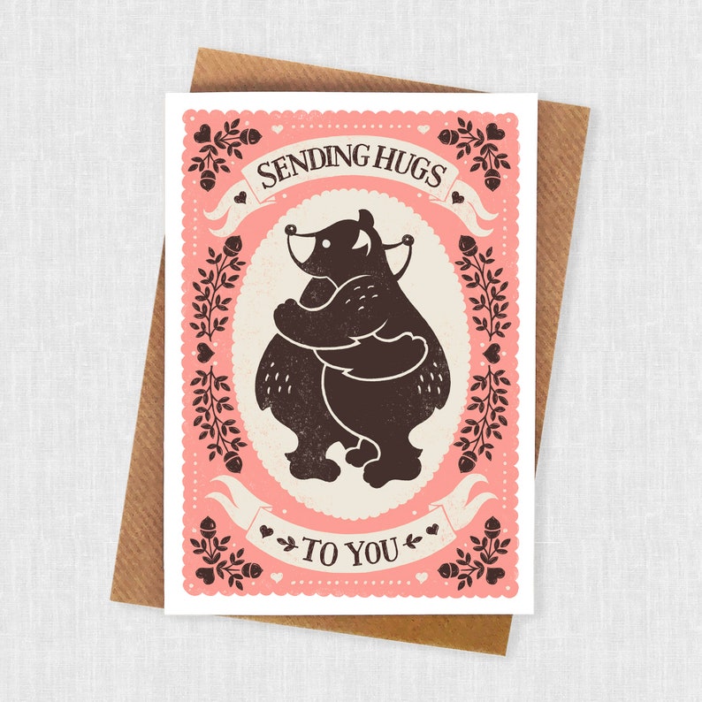 May include: A pink card with a black silhouette of two bears hugging. The card has a white border and says "Sending Hugs To You" in a vintage font.