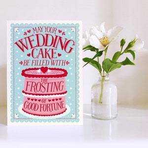 May include: A light blue card with a white scalloped border. The card features a pink and white wedding cake with the text "May your wedding cake be filled with the frosting of good fortune".