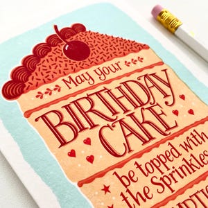 May include: A birthday card with a red and white cake illustration. The card says "May your birthday cake be topped with the sprinkles".