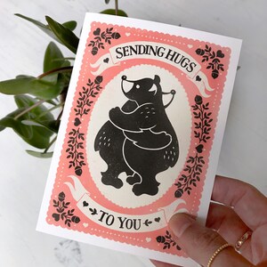 May include: A pink and white card with a black silhouette of two bears hugging. The card says "Sending Hugs To You" with hearts and floral designs.