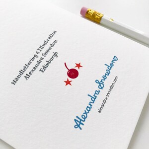 May include: A white card with blue text that reads "Handlettering & Illustration Alexandra Snowdon Edinburgh" and a red cherry with two red stars. The text "Alexandra Snowdon" is written in blue cursive with a heart at the end. Below the cursive text is the website address "alexandra-snowdon.com".