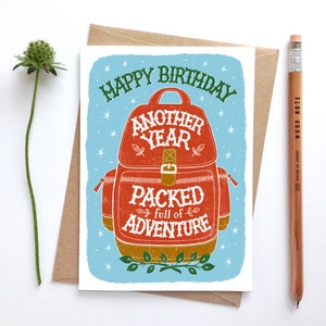 May include: Happy birthday card with a red and brown backpack illustration. The backpack has the text "Another Year Packed full of Adventure" on it.