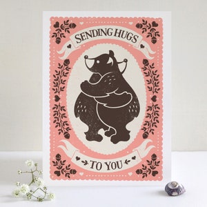 May include: A pink card with a white border and a brown illustration of two bears hugging. The card says "Sending Hugs To You" in a brown font.