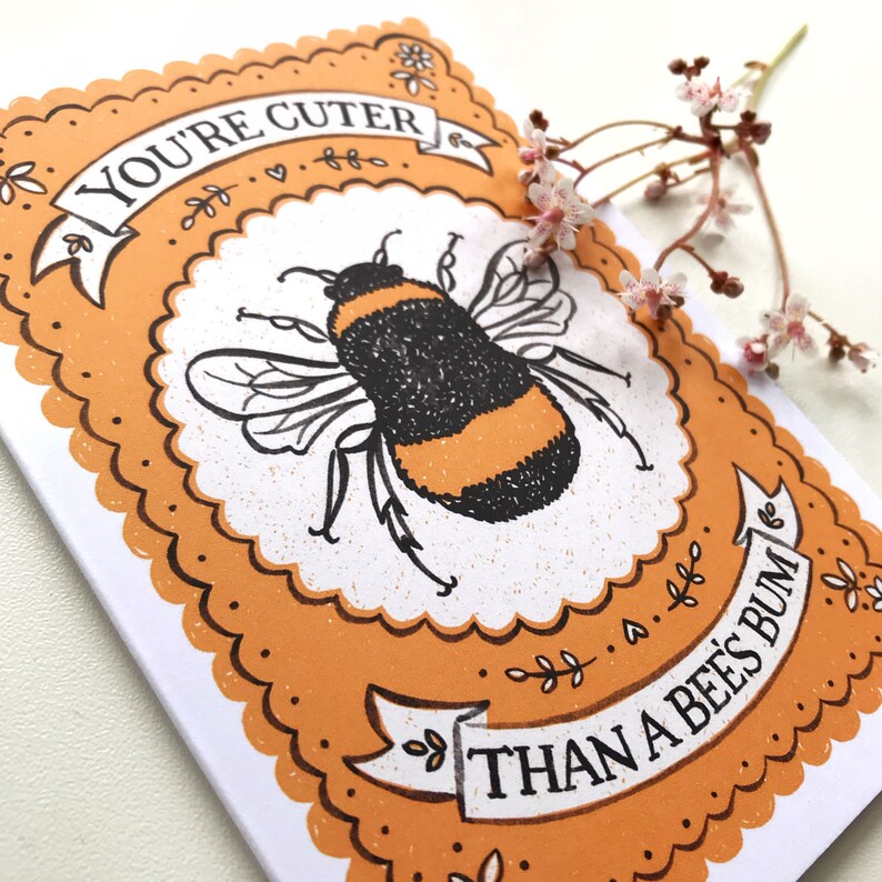 May include: A greeting card with an orange background and a black and yellow bumblebee illustration. The card has a white border and the text "You're cuter than a bee's bum." in a brown banner.