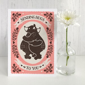 May include: A pink card with a black and white illustration of two bears hugging. The card says "Sending Hugs To You" with hearts and other decorative elements.
