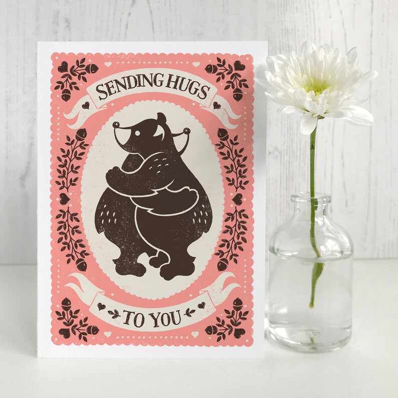 May include: A pink card with a black and white illustration of two bears hugging. The card says "Sending Hugs To You" with hearts and other decorative elements.