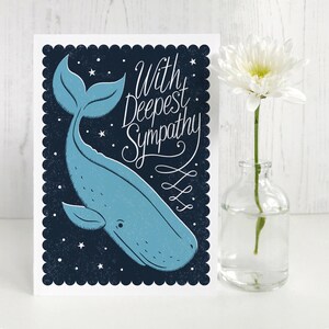 May include: A sympathy card with a blue whale illustration on a dark blue background with white stars. The card has the words "With Deepest Sympathy" in script. A white flower in a small glass vase is to the right.