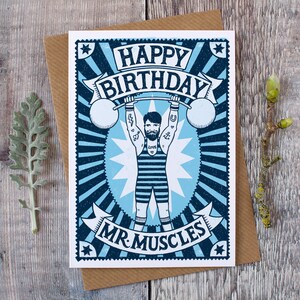 Pu&ograve; includere: Un biglietto di auguri di compleanno blu e bianco con un'illustrazione di un uomo forte che solleva pesi. Il biglietto dice "Buon compleanno Mr. Muscles".
