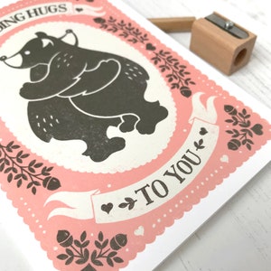 May include: A pink greeting card with a black and white illustration of two bears hugging. The card has a white banner with the text "Sending Hugs to You".