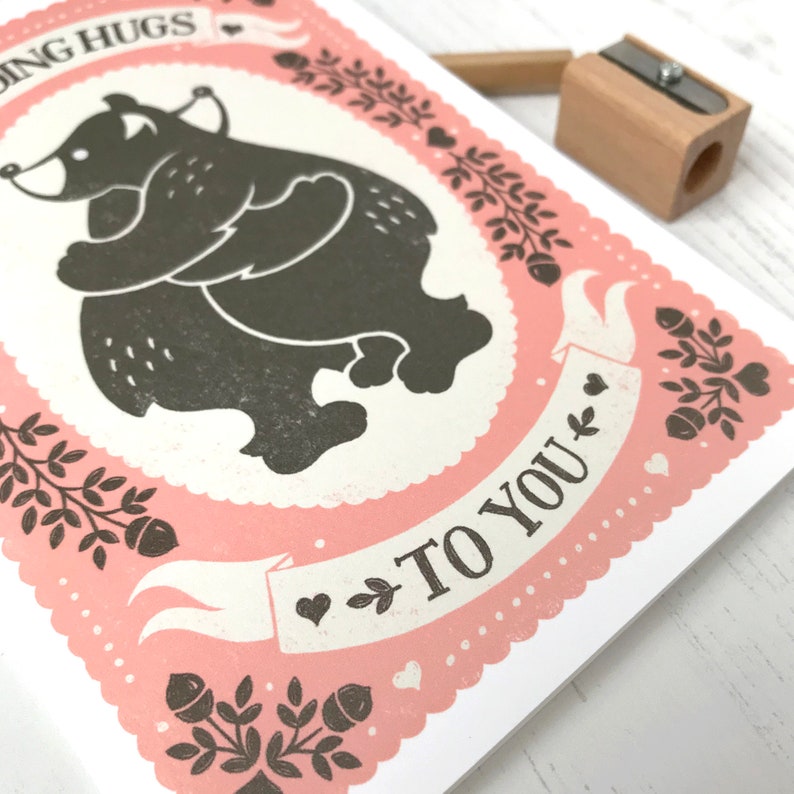 May include: A pink greeting card with a black and white illustration of two bears hugging. The card has a white banner with the text "Sending Hugs to You".