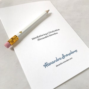 Pu&ograve; includere: Un biglietto bianco con il testo "Handlettering & Illustration Alexandra Snowden" in blu e un cuore dorato. Il testo "Alexandra Snowden" &egrave; in blu con un cuore dorato e il testo "alexandra-snowdon.com" in nero.