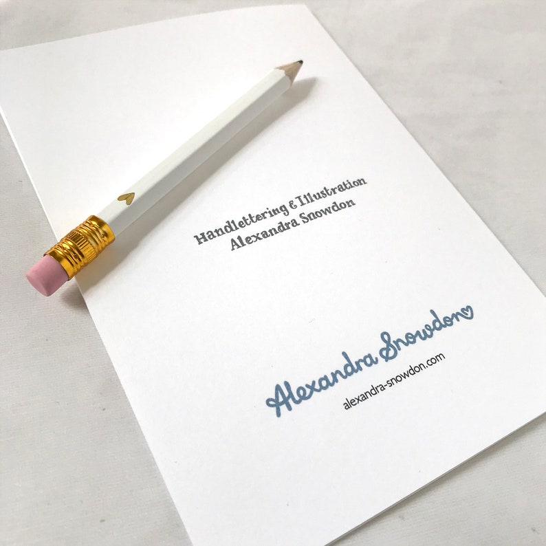 Pu&ograve; includere: Un biglietto bianco con il testo "Handlettering & Illustration Alexandra Snowden" in blu e un cuore dorato. Il testo "Alexandra Snowden" &egrave; in blu con un cuore dorato e il testo "alexandra-snowdon.com" in nero.