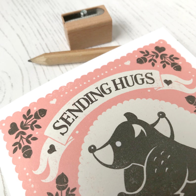 May include: A pink and white greeting card with a black and white illustration of a bear. The card says "Sending Hugs" in a banner.