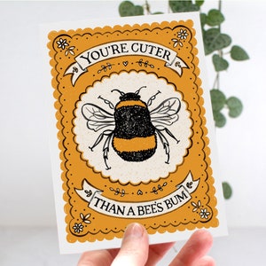 May include: A yellow and white card with a black and yellow bumblebee illustration. The card has a scalloped border and the text "You're cuter than a bee's bum".