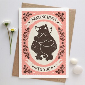 May include: A pink and white greeting card with a black silhouette of two bears hugging. The card says "Sending Hugs To You" in a decorative font.