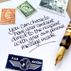 May include: A white paper with handwritten text and vintage stamps. The text says, "You can choose to have your card sent direct to the recipient with your own personal message inside." A fountain pen is visible. Stamps are also present.