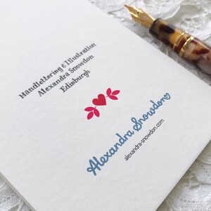 May include: A white card with the text "Handlettering & Illustration Alexandra Snowdon Edinburgh" and a blue handwritten logo that says "Alexandra Snowdon" with a website address below it. The logo is accompanied by two pink stylized leaves and a red heart.