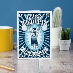 Pu&ograve; includere: Un biglietto di auguri di compleanno blu e bianco con un'illustrazione di un uomo che solleva pesi con il testo "Happy Birthday Mr. Muscles".
