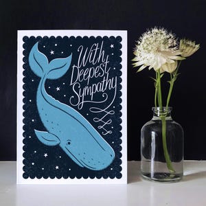 May include: A sympathy card featuring a blue whale illustration on a dark blue background with stars. The card reads "With Deepest Sympathy" in white script. A clear glass vase with white flowers is to the right.